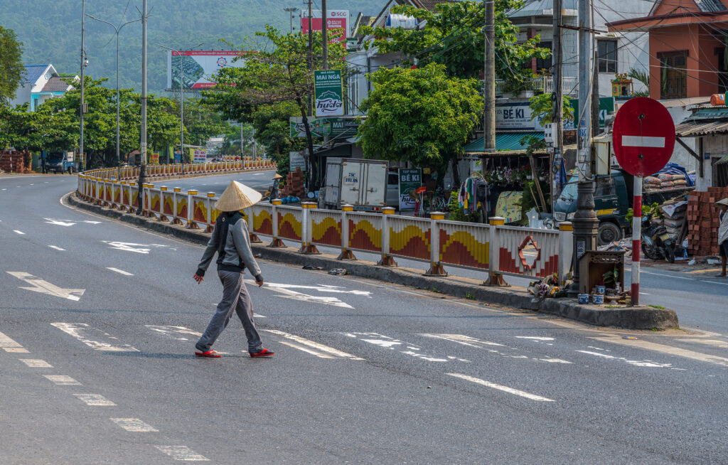 A woman wearing a conical hat and red shoes crosses a broad road in central Vietnam