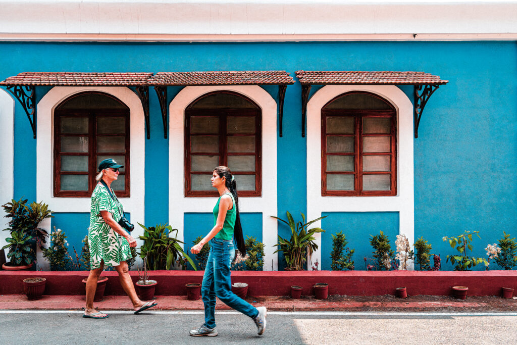 Two women pass one another in front of a turquoise house with white window surrounds.