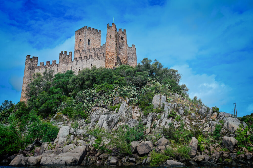 Medieval Almourol Castle rises above a rocky, tree-covered island in the Tagus River beneath a dramatic blue sky in central Portugal.